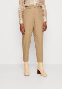 Pantalons cargo beiges avec une coupe fuselée, présentant des poches latérales et une fermeture par bouton. Assortis à des bottines à talons légers beiges.