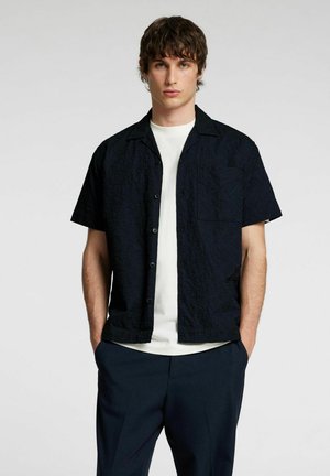 Chemise noire à manches courtes en tissu texturé, avec un boutonnage à l'avant et une poche poitrine unique, portée par-dessus un T-shirt blanc à col rond.