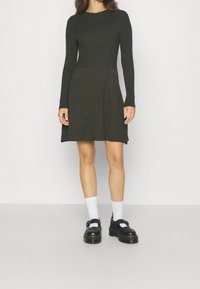 Mörkgrön ribbad stickad klänning med lång ärm, figurnära bodice, utsvängd kjol och sidoklaffar. Matchad med vita strumpor och svarta skor.