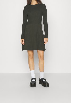 Abito a maglia a coste verde scuro con maniche lunghe, corsetto aderente, gonna svasata e spacchi laterali. Abbinato a calze bianche e scarpe nere.