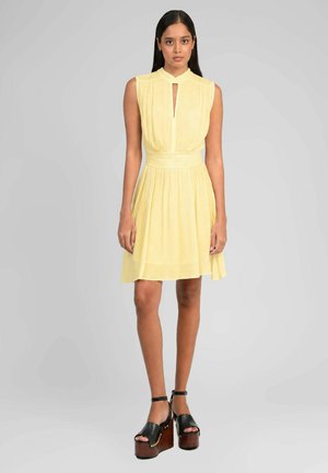 Jeune femme debout portant une robe jaune sans manches jusqu'aux genoux et des sandales à plateforme noires sur un fond gris uni.