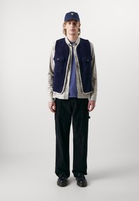 Jūras spēku flīsa veste ar divām priekšējām kabatām uz pārbaudītas kreklu, apvienota ar melniem biksēm un melnām kurpēm, virsū valkāta jūras spēku cepure.