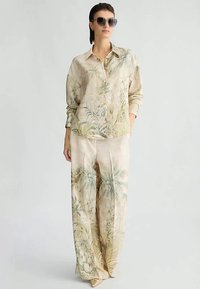 Donna che indossa una camicia a bottoni beige con stampa tropicale e pantaloni larghi coordinati, accessoriata con occhiali da sole grandi e scuri.