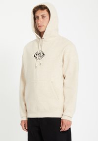 Beige hoodie met een voorzak, een trekkoordkap en een zwart logo op de borst. Zachte textuur.