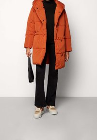 Orange pufferväst med huva, med ett kviltat mönster och svarta knappar. Bärs över en svart topp med vida svarta byxor och sneakers.