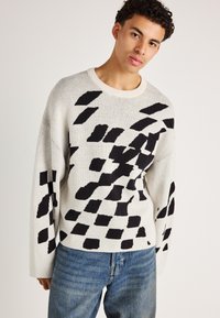 Oversize genser med rund hals i lys grå, med sterke svarte grafiske mønstre. Myk strikket tekstur med drop shoulder-design og ribbede mansjetter.