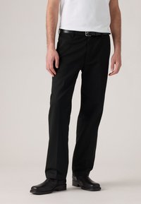 Man wearing black straight-leg trousers, black leather belt, black dress shoes, and a white t-shirt, standing on a light gray background.