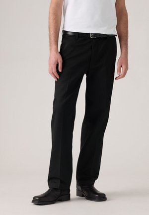 Man in zwarte recht-toe-rechtdoor broek, zwarte leren riem, zwarte nette schoenen en een wit T-shirt, staand tegen een lichtgrijze achtergrond.