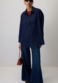 Camicia blu navy con vestibilità rilassata, indossata sopra un top bordeaux, abbinata a jeans in denim a gamba larga e una piccola borsa marrone.