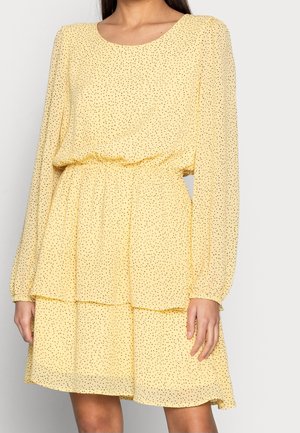 Femme portant une robe jaune à manches longues avec de petits pois noirs, avec une taille cintrée et une jupe à volants.