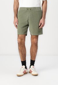 Grüne Cordshorts mit einem elastischen Bund, Kordelzug und zwei seitlichen Taschen. Getragen mit weißen Sneakers mit roten Akzenten.