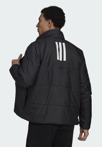 Giacca a vento nera con colletto alto, texture trapuntata e tre strisce bianche sul retro. Chiusura con zip e tasche laterali.