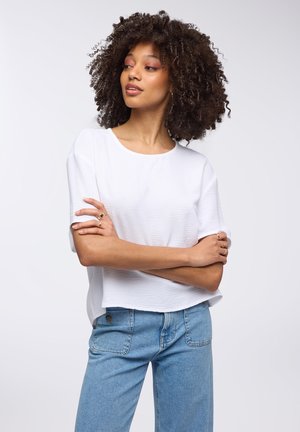 Top bianco a vita alta con un leggero motivo testurizzato, abbinato a jeans denim blu chiaro. La modella ha capelli ricci, le braccia incrociate, ed è leggermente di lato.