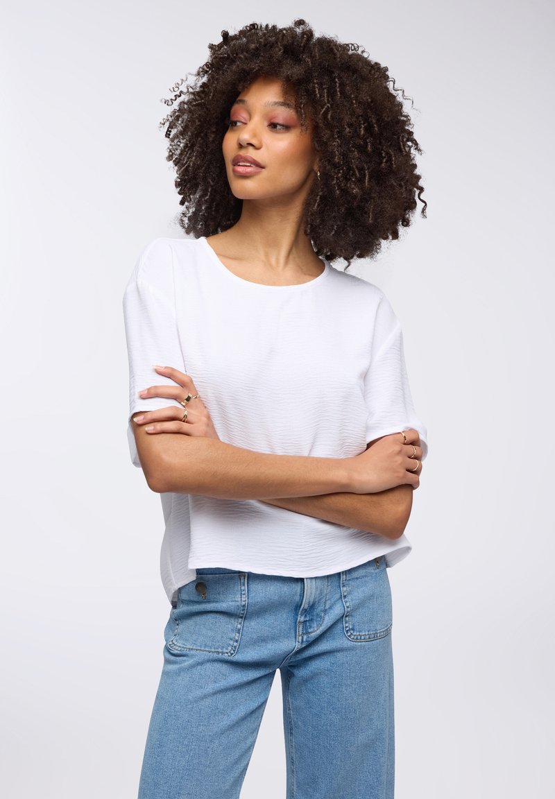Top bianco a vita alta con un leggero motivo testurizzato, abbinato a jeans denim blu chiaro. La modella ha capelli ricci, le braccia incrociate, ed è leggermente di lato.
