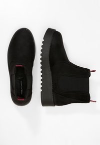 Musta mokkanahka Chelsea-saappaat paksuilla uritelluilla pohjilla, elastisilla sivupaneeleilla ja punaisilla vetonauhoilla valkoista taustaa vasten.