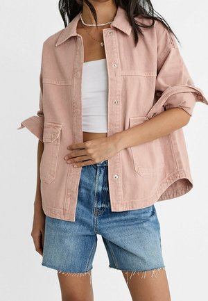 Kobieta nosząca oversize różową kurtkę zapinaną na guziki z podwiniętymi rękawami, biały krótki top, niebieskie jeansowe szorty i warstwowe naszyjniki.