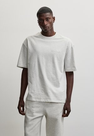 Mand stående med fronten mod kamera iført lysegrå oversized T-shirt og matchende joggingbukser mod en ensfarvet lys baggrund.