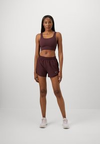 Bordeauxfarbenes Sport-BH- und Shorts-Set. Der BH hat breite Träger; die Shorts haben einen elastischen Bund und das silberne Nike-Logo. Die Sneakers sind hellrosa.