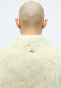 Krämfärgad fluffig kappa med en texturerad yta, prydd med en liten silver-emblem i mitten av ryggen, designad med en avslappnad silhuett.