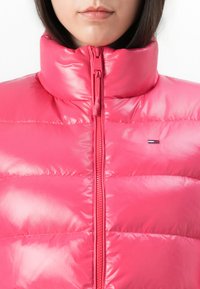 Kadar izbliza osobe koja nosi sjajnu ružičastu puffer jaknu s visokim ovratnikom i prednjim zatvaračem, prikazujući dio njihovih usana i brade.