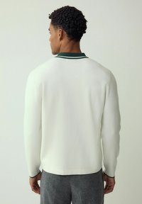 White sweater with green collar accents, long sleeves, and a straight hem, shown from the back, textured fabric, paired with gray pants.