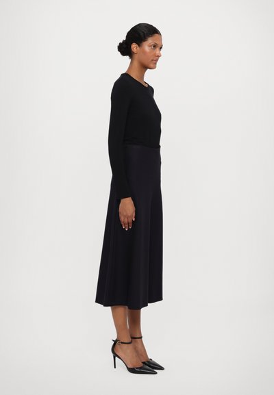 Top negro de manga larga combinado con una falda oscura y fluida. La modelo lleva tacones negros puntiagudos, mostrando una silueta suave y elegante.