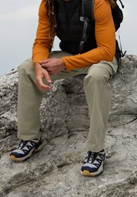 Buty do chodzenia po górach mają czarny, szary i niebieski design z mocną podeszwą. Osoba nosi beżowe spodnie i pomarańczową bluzę z długim rękawem.