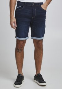 Shorts en denim indigo foncé avec ourlets retroussés, dotés de poches latérales et d'une coupe slim. Associés à des baskets noires sur un fond neutre.