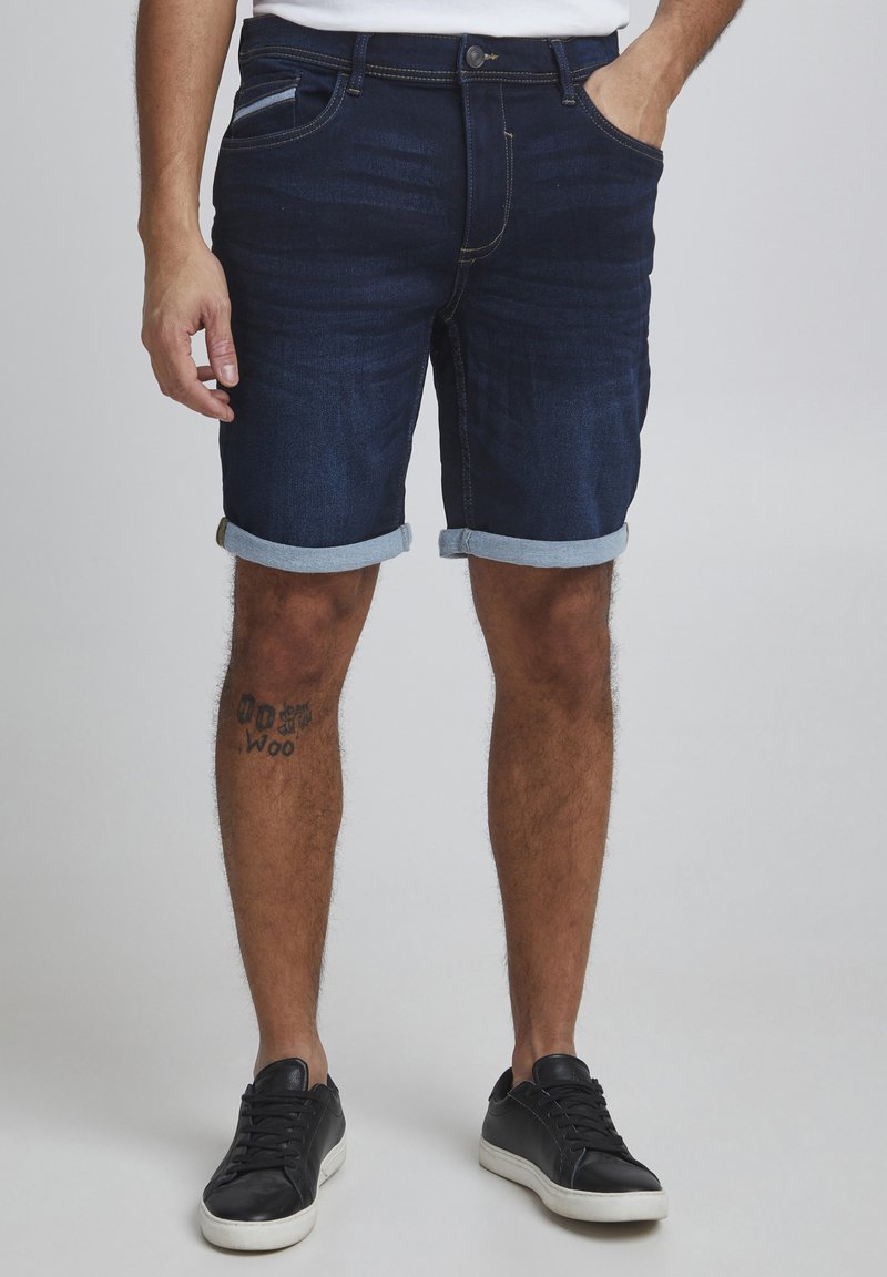 Shorts en denim indigo foncé avec ourlets retroussés, dotés de poches latérales et d'une coupe slim. Associés à des baskets noires sur un fond neutre.