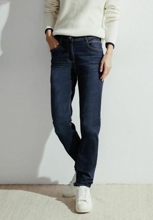 Dark blue denim jeans with a straight fit, featuring contrast stitching, five pockets, and a zip fly. Paired with a white knit sweater.