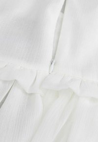 Biała sukienka z teksturowanej tkaniny, z falbanami wzdłuż dołu i zapięciem na zamek. Miły, lekki materiał.