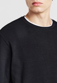 Felpa nera con texture, colletto tondo e un collo interno bianco. Presenta un design aderente e polsini a coste.