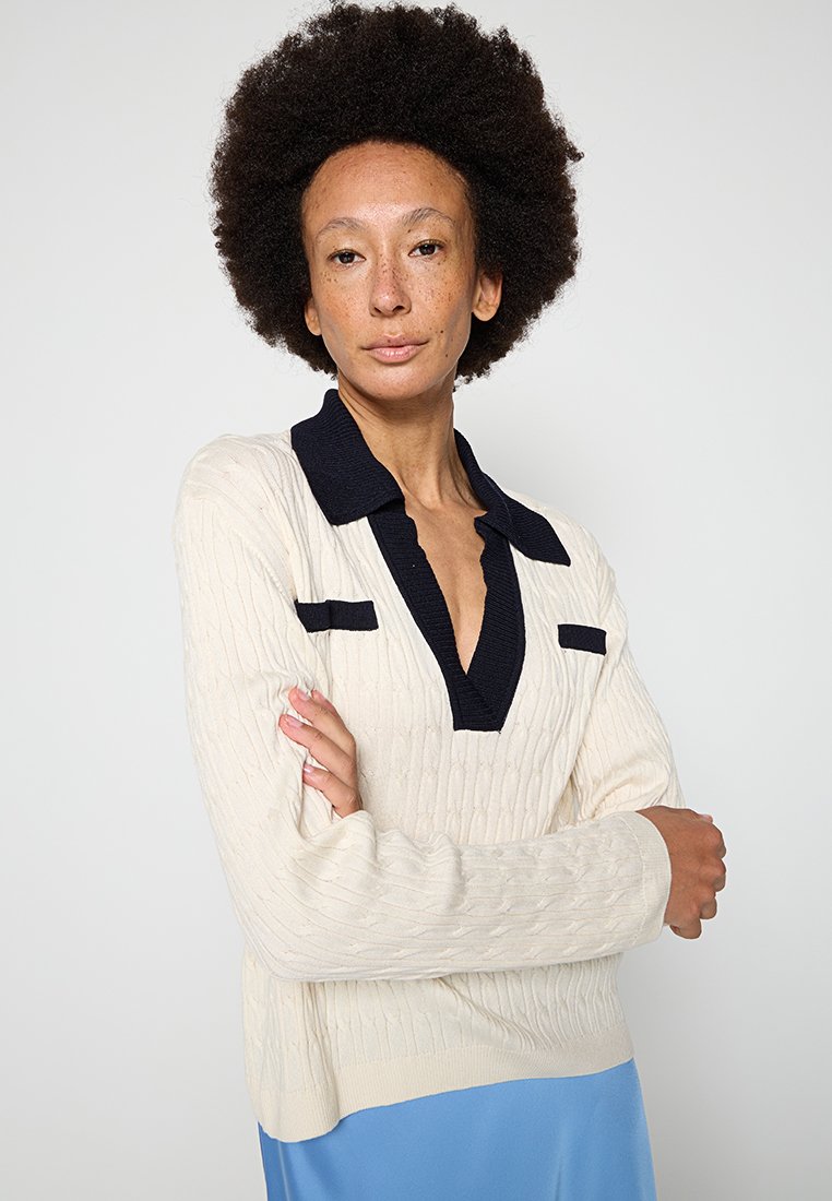 Pullover a maglia cable color crema con colletto blu navy e dettagli sulle tasche. Presenta maniche lunghe e una vestibilità comoda. Texture morbida.