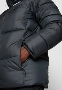 Chaqueta de plumas en azul marino oscuro, con cierre frontal de cremallera, relleno acolchado grueso y un bolsillo lateral con detalle de cierre elástico.