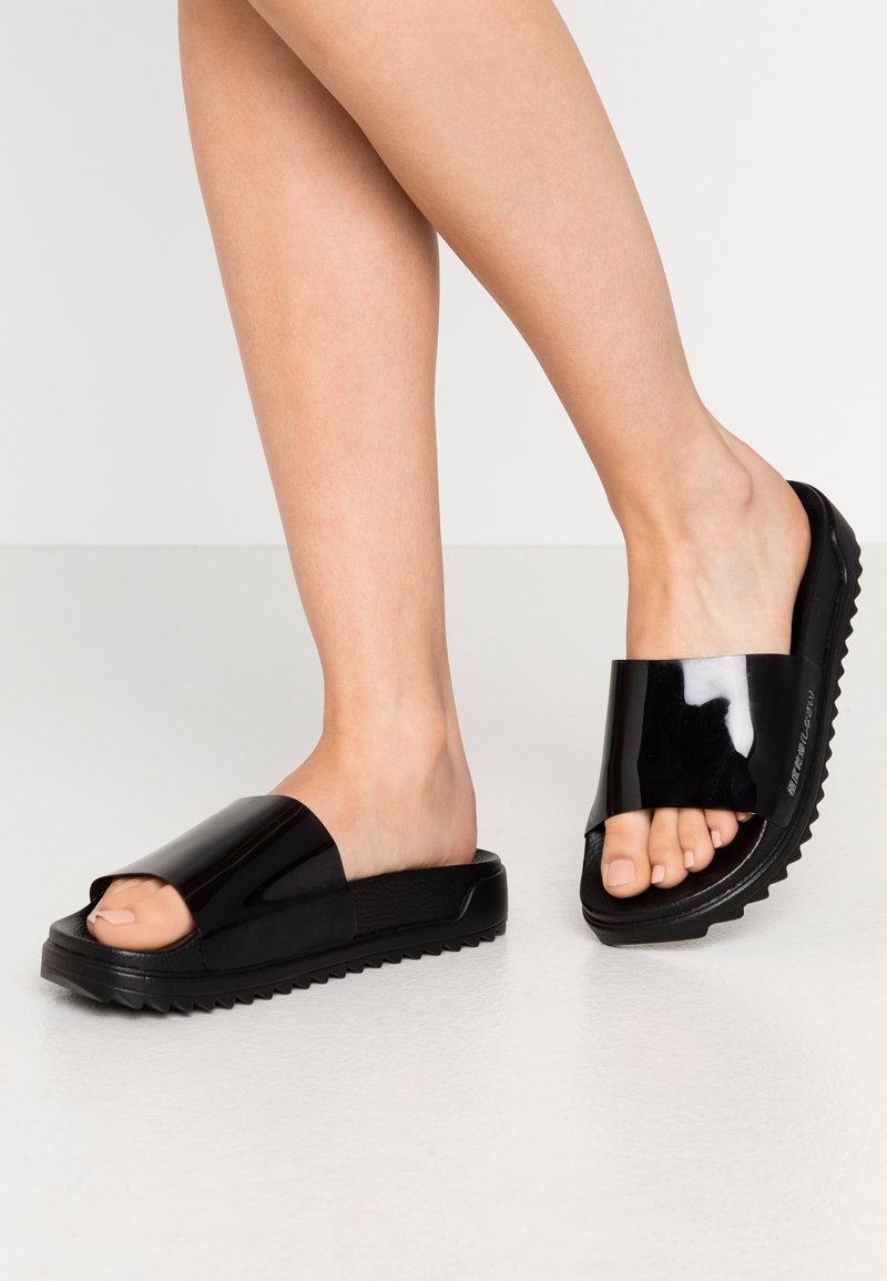 Sorte slidesandaler med en blank overflade, åben tå og rillet sål. Glat tekstur, bred rem over foden, minimalistisk design.