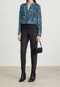 Chemisier en soie bleu et vert à motifs associé à un pantalon noir ajusté et des bottines noires à talons; tenant un petit sac à main noir.