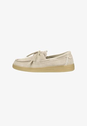 Beige suède mocassinstijl schoen met vetersluiting en gestructureerde lichtbruine rubberen zool, zijaanzicht op een witte achtergrond.
