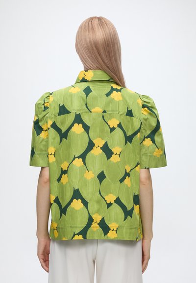 Personne blonde de dos portant une chemise verte à manches courtes avec un grand motif feuillu et floral jaune, et un pantalon blanc.