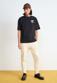 Black short-sleeve T-shirt with white graphic, paired with light beige joggers. Wearing a brown cap and black shoes with a patterned design.