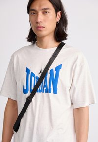 Svijetlosiva oversized majica s velikim plavim natpisom "JORDAN" i crnim logotipom. Akcentirana crnom trakom preko ramena.