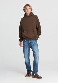 Felpa con cappuccio marrone e tasca frontale, abbinata a jeans azzurri chiari e scarpe marroni con suola a strati e cuciture decorative.