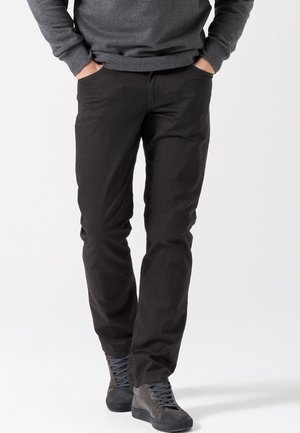 Mężczyzna ubrany w czarne spodnie, szarą koszulę z długim rękawem i ciemnoszare wysokie trampki, z rękami w kieszeniach, stoi na białym tle.