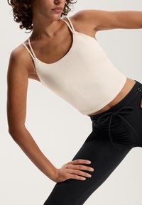 Világos bézs színű, rövidített tank top vékony pántokkal, fekete, magas derekú leggings-szel párosítva, sima textúrával és testhez simuló fazonnal.