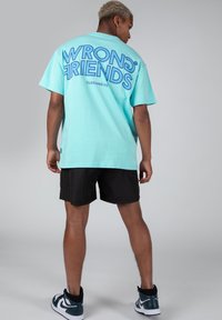 Camiseta oversized turquesa con gran texto azul "AMIGOS INCORRECTOS" en la parte de atrás, combinada con pantalones cortos negros y zapatillas negras/verdes.