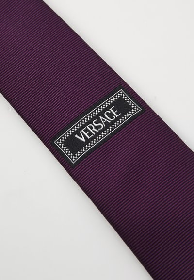 Lila selyem nyakkendő bordázott mintával, fekete téglalap alakú címkével, fehér felirattal. A címkén a "VERSACE" felirat díszes keretben található.