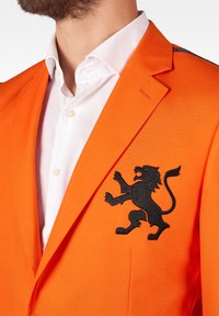 Oranž blazer musta lõviga tikandiga vasakul rinnal, narmastatud krae ja valge särk all. Sile kangast tekstuur.
