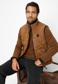 Gilet marrone trapuntato con colletto alto, bordi neri e tasca frontale. Indossato sopra un maglione marrone e abbinato a jeans neri, seduto su una sedia.