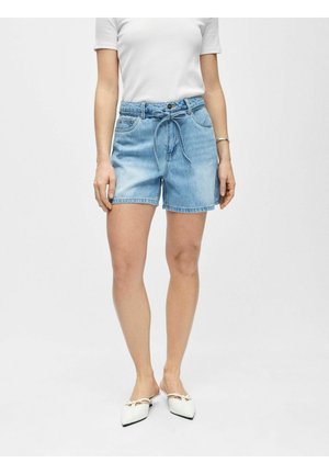 Femme portant un short en jean bleu clair avec une ceinture nouée, une chemise blanche à manches courtes, des ballerines blanches à bout pointu et un bracelet en argent.