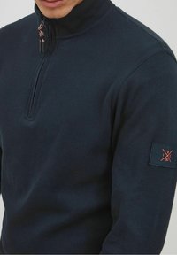 Navy-Half-Zip-Pullover aus geripptem Stoff. Verfügt über einen hohen Kragen, eine dekorative Kordel und ein Patch mit orangefarbenen Akzenten auf dem Ärmel.