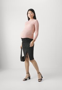 Schwangerschaftsoutfit mit einem strukturierten rosa Langarmtop und einem schwarzen, gerafften Rock mit einem Schlitz vorne. Kombiniert mit schwarzen, geschnallten Highheels.